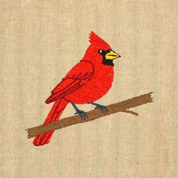 Embroidered cardinal bird design on a beige fabric background, perched on a brown branch with red, yellow, black, and blue-gray details.