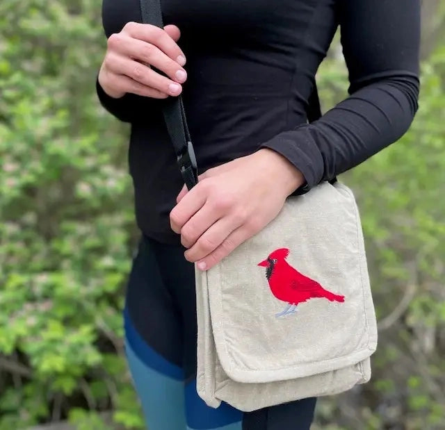 A person outdoors wearing a beige canvas crossbody bag with a red cardinal bird embroidery on the front flap, paired with a black long-sleeve top.