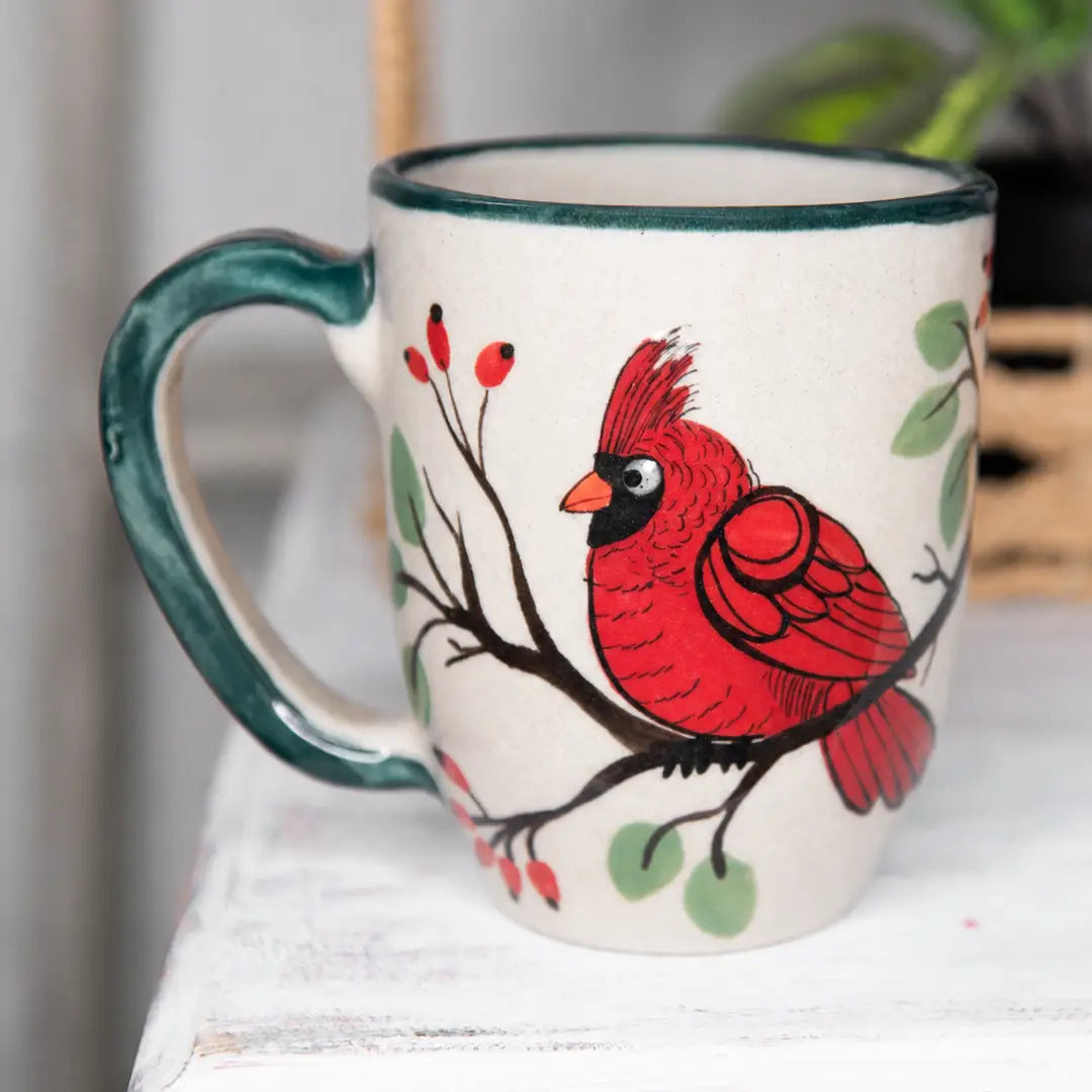 A ceramic mug with a cream-colored body, featuring a hand-painted red cardinal perched on a branch with green leaves and red berries. The mug has a dark green handle and rim, and is shown resting on a white wooden surface.