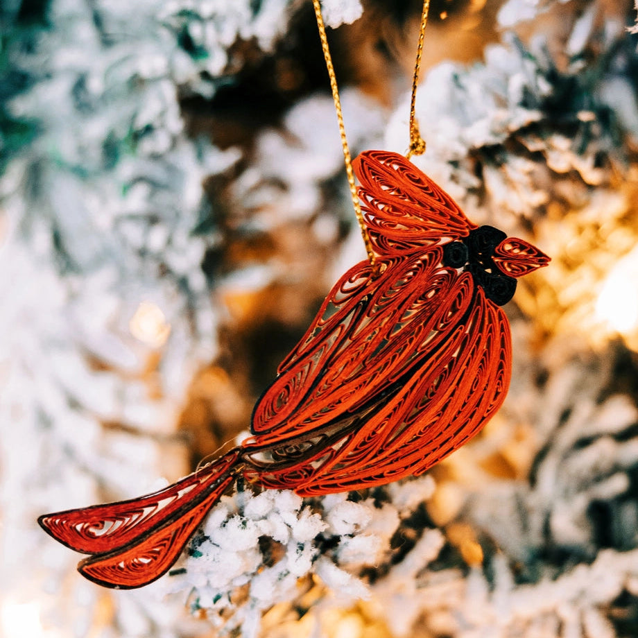 A red cardinal Christmas ornament with a gold hanging string, photographed hanging against a blurred frosted tree background with warm lights.