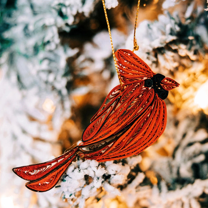 A red cardinal Christmas ornament with a gold hanging string, photographed hanging against a blurred frosted tree background with warm lights.