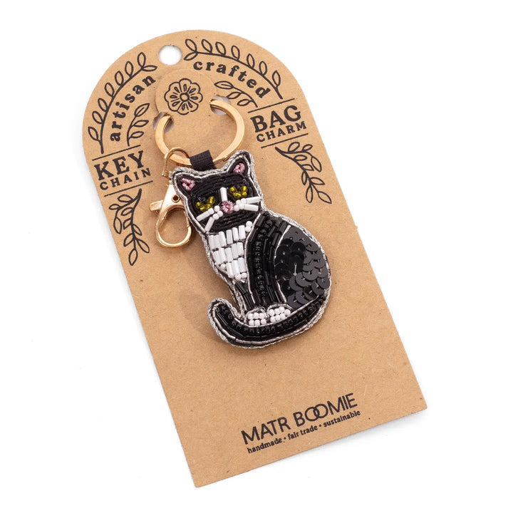 A black and white cat-shaped keychain charm with yellow eyes and a gold clasp, attached to a brown kraft paper display card labeled 'Matr Bodme'.