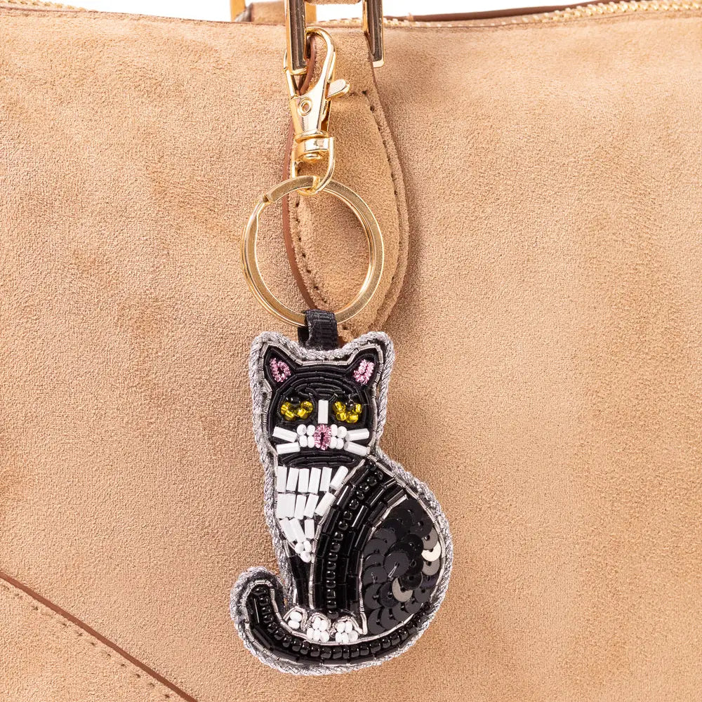 A black cat-shaped keychain charm with sequins and embroidered details, hanging from a gold ring on a beige suede handbag.