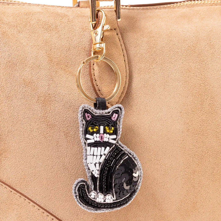 A black cat-shaped keychain charm with sequins and embroidered details, hanging from a gold ring on a beige suede handbag.
