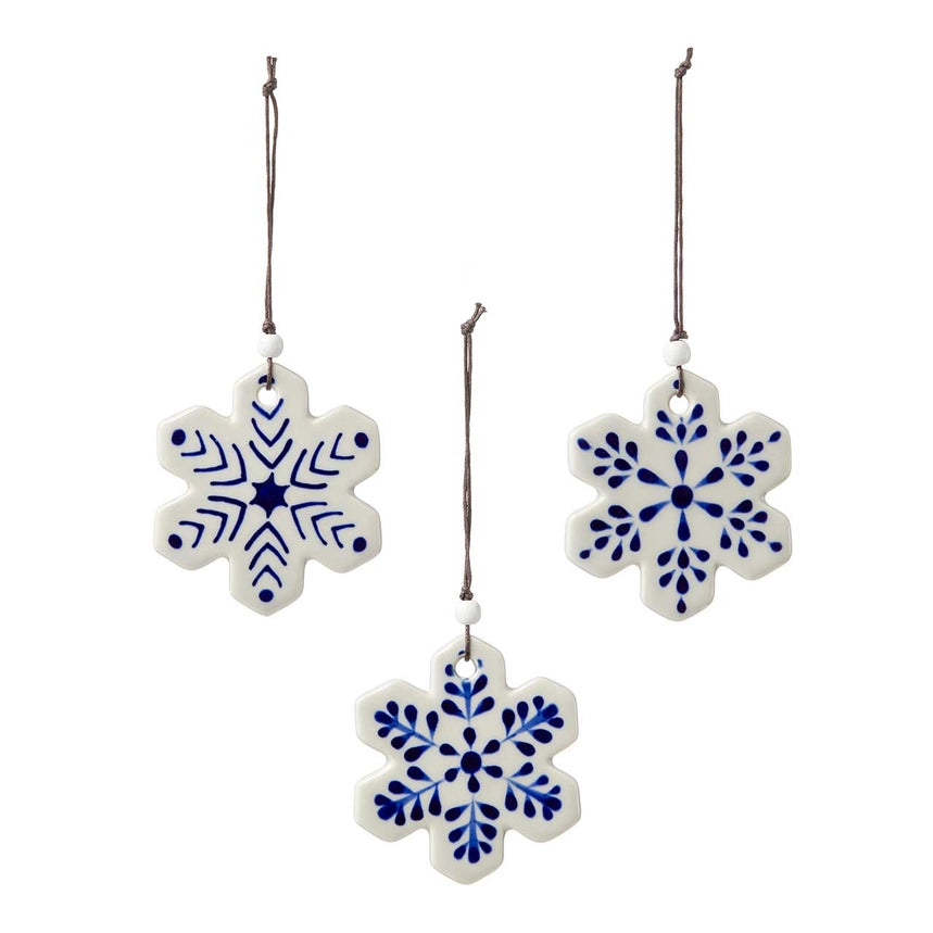 Three ceramic snowflake ornaments in white with blue hand-painted patterns, hanging from brown strings against a plain white background.