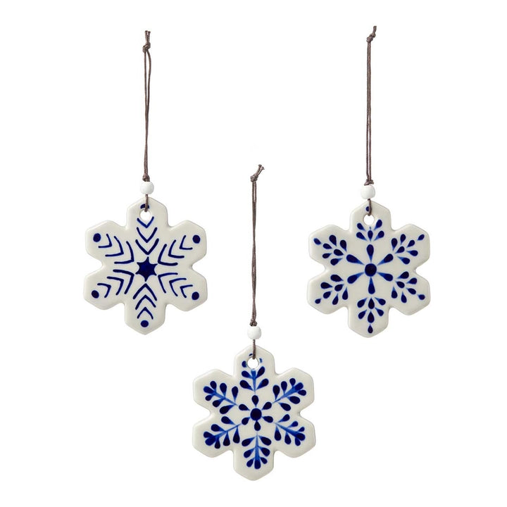 Three ceramic snowflake ornaments in white with blue hand-painted patterns, hanging from brown strings against a plain white background.
