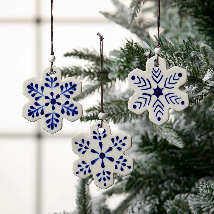 Three white ceramic snowflake Christmas ornaments with blue hand-painted patterns hanging from a snowy evergreen tree.