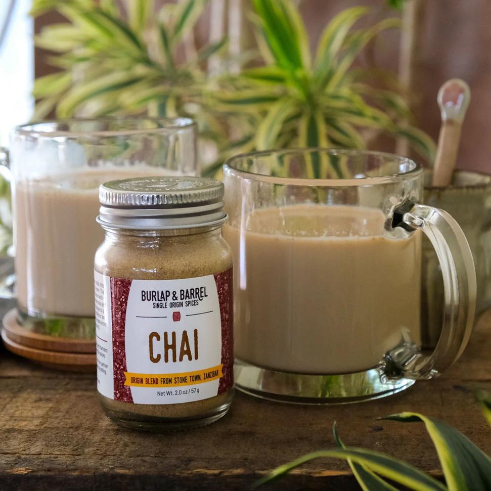 Jar of Burlap & Barrel Chai spice blend with a glass mug of chai tea on a wooden surface.