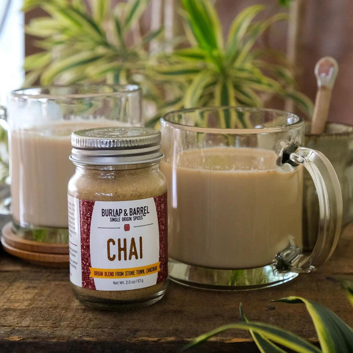 Jar of Burlap & Barrel Chai spice blend with a glass mug of chai tea on a wooden surface.