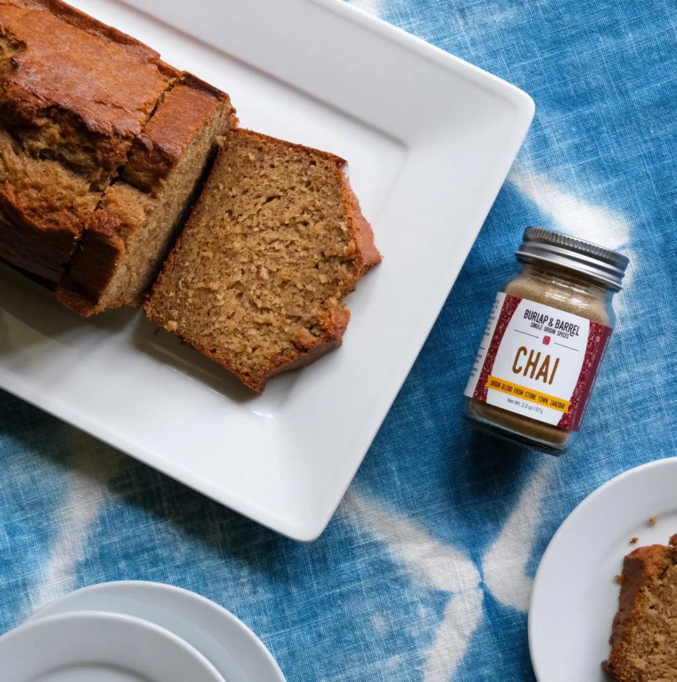 Loaf of banana bread on a white plate with a jar of Chai spice mix on a blue textured surface.
