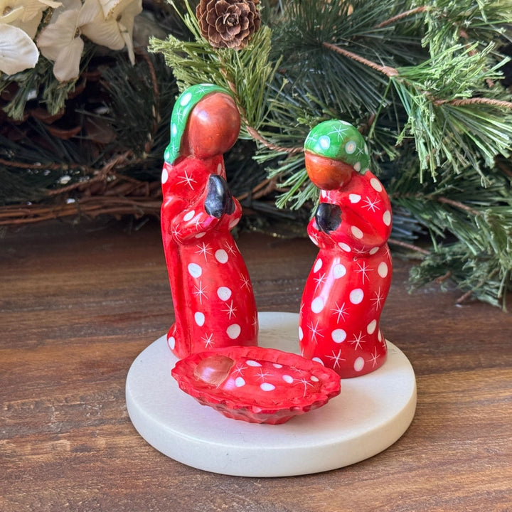A small Christmas figurine set on a round white base, featuring two red-robed figures with green head coverings and white polka dots, standing beside a matching red bowl. The figurine is photographed on a wooden surface with pine branches in the background.