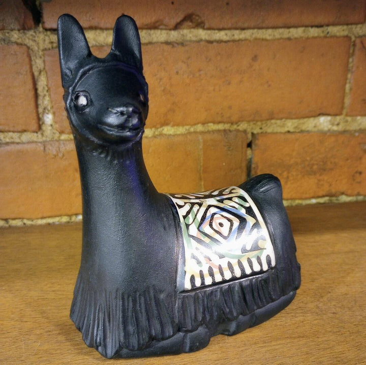 Black decorative llama figurine with a patterned saddle on a wooden surface with a brick wall background.