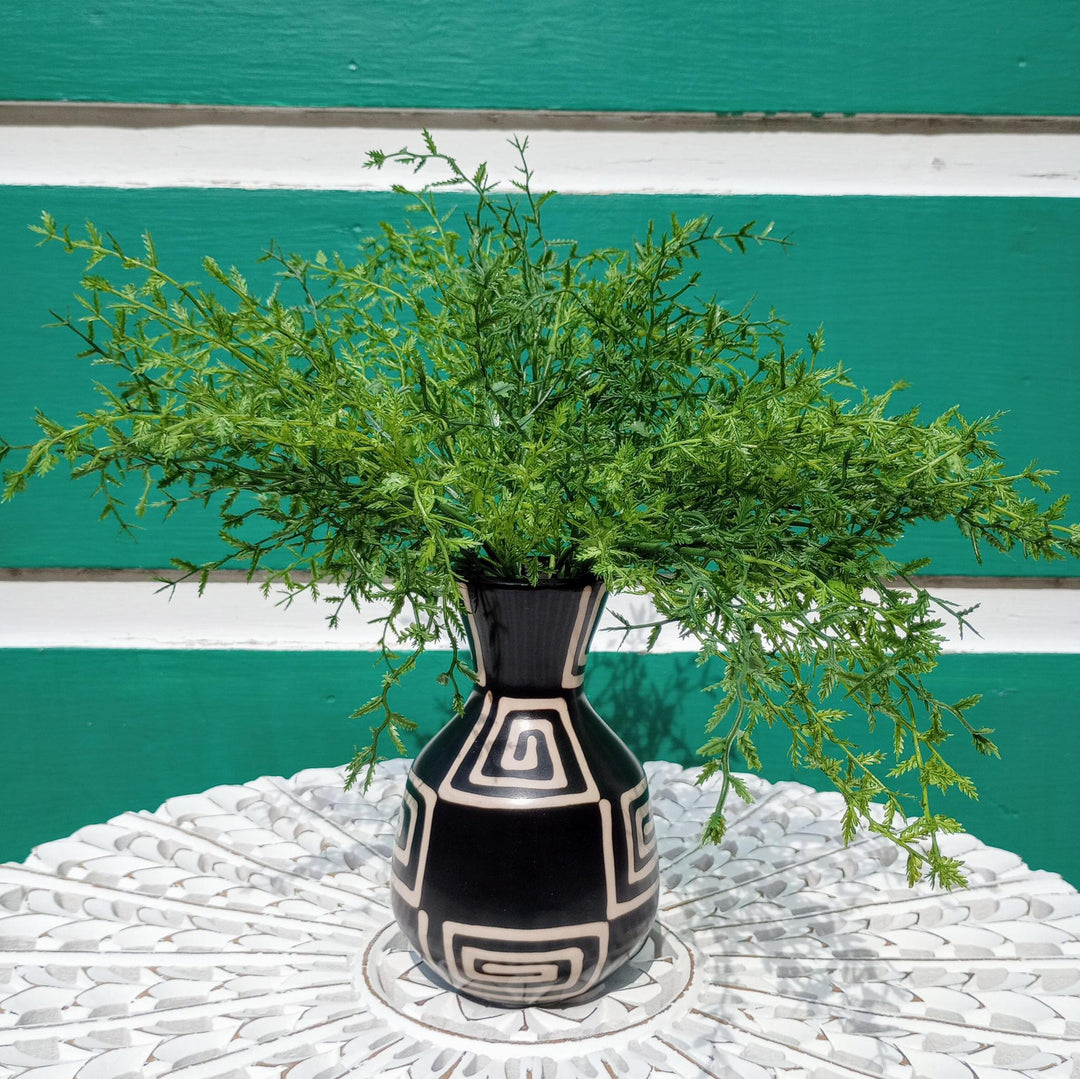 Black vase with white patterns holding green branches against a teal and white striped background