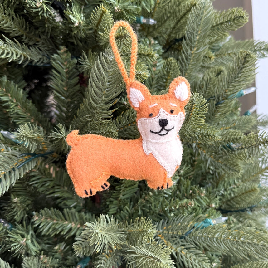 A felt corgi dog Christmas ornament hanging from a green Christmas tree branch. The ornament is made of orange and cream felt with black embroidered facial features and a hanging loop.
