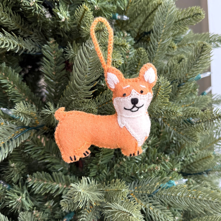 A felt corgi dog Christmas ornament hanging from a green Christmas tree branch. The ornament is made of orange and cream felt with black embroidered facial features and a hanging loop.