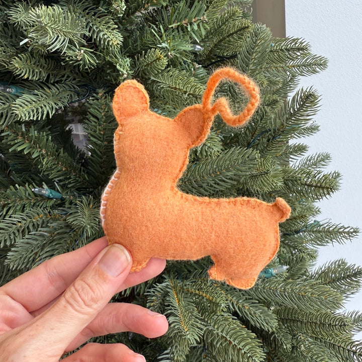 A hand holds a small felt Christmas ornament shaped like a dog in orange-brown, positioned in front of green Christmas tree branches.