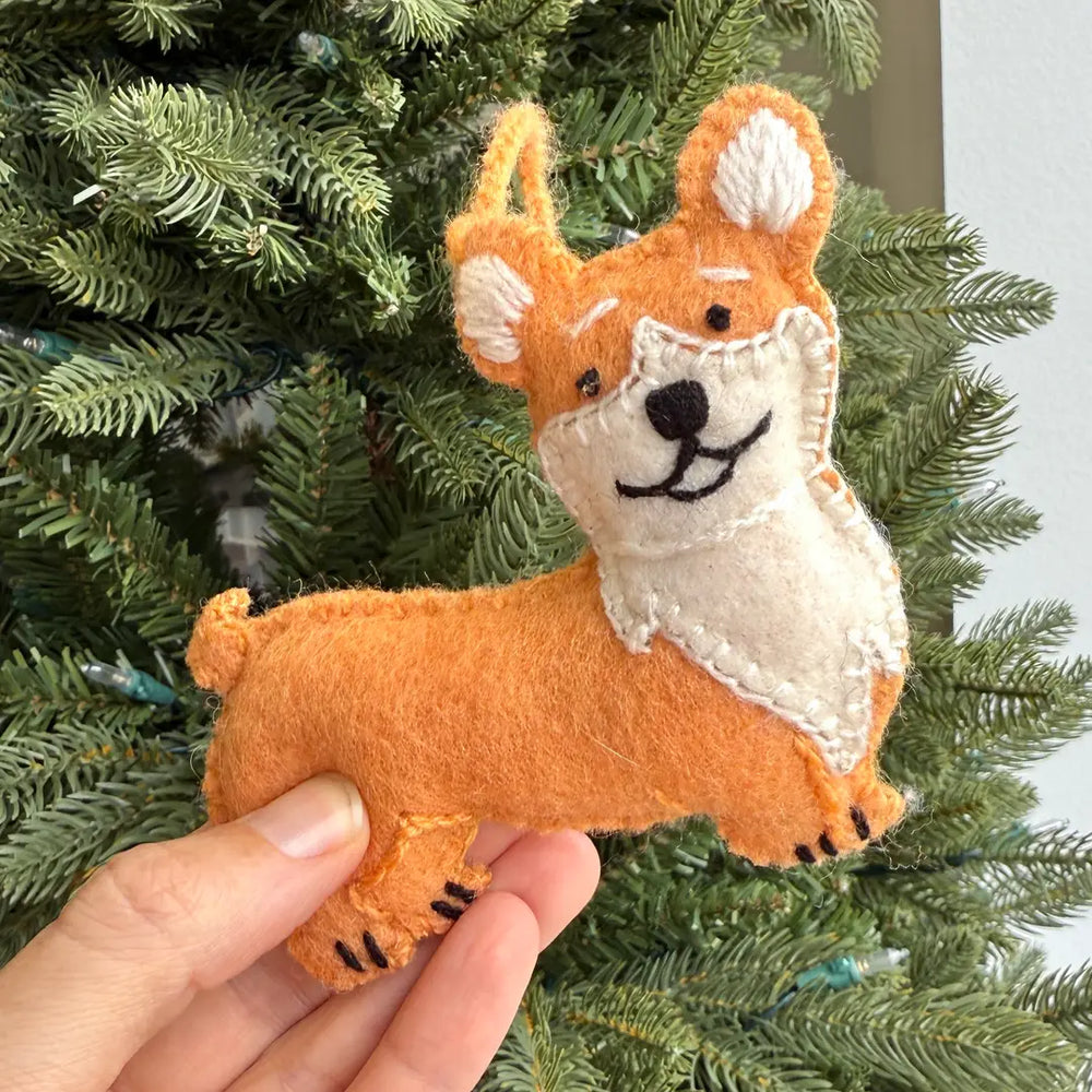 A hand holds a felt Corgi Christmas ornament in front of a green Christmas tree. The ornament is made from orange and cream felt with black embroidered facial features.