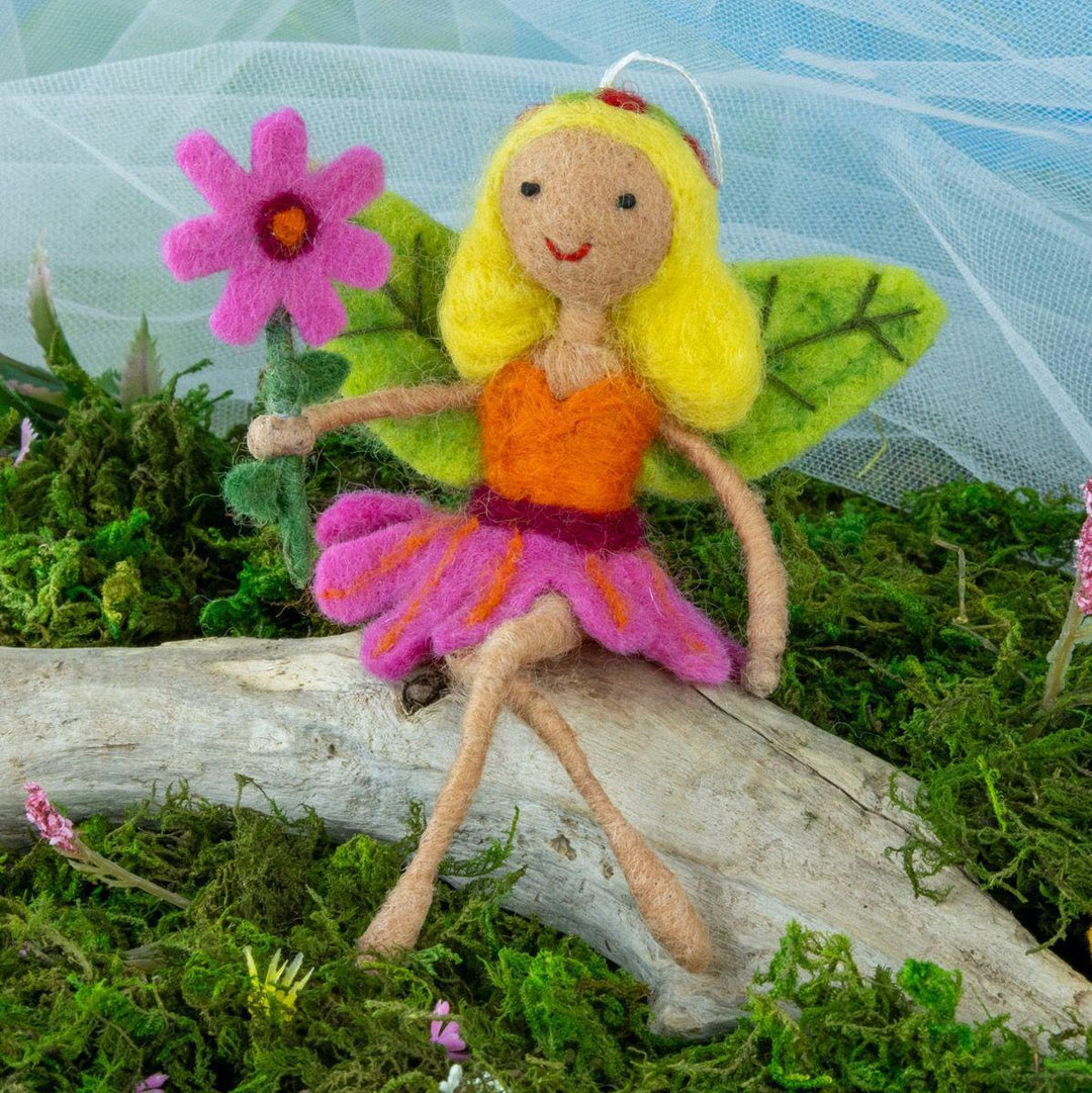 Felt fairy ornament with yellow hair and orange dress sitting on a log with a pink flower in a green, natural setting.