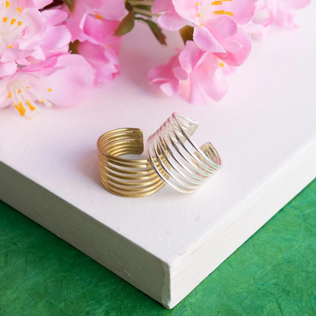 Two spiral-shaped rings, one gold and one silver, on a white surface with pink flowers in the background.