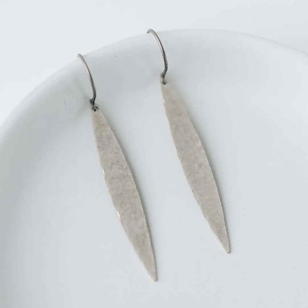 A pair of minimalist silver dangle earrings with textured, hammered elongated drop shapes and simple fishhook wires, displayed on a clean white surface.