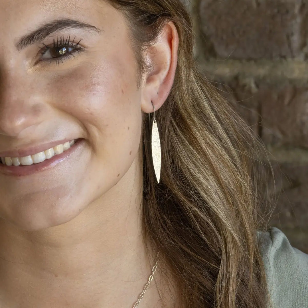 A close-up of a smiling woman wearing a single long, feather-shaped gold dangle earring against a blurred brick wall background.