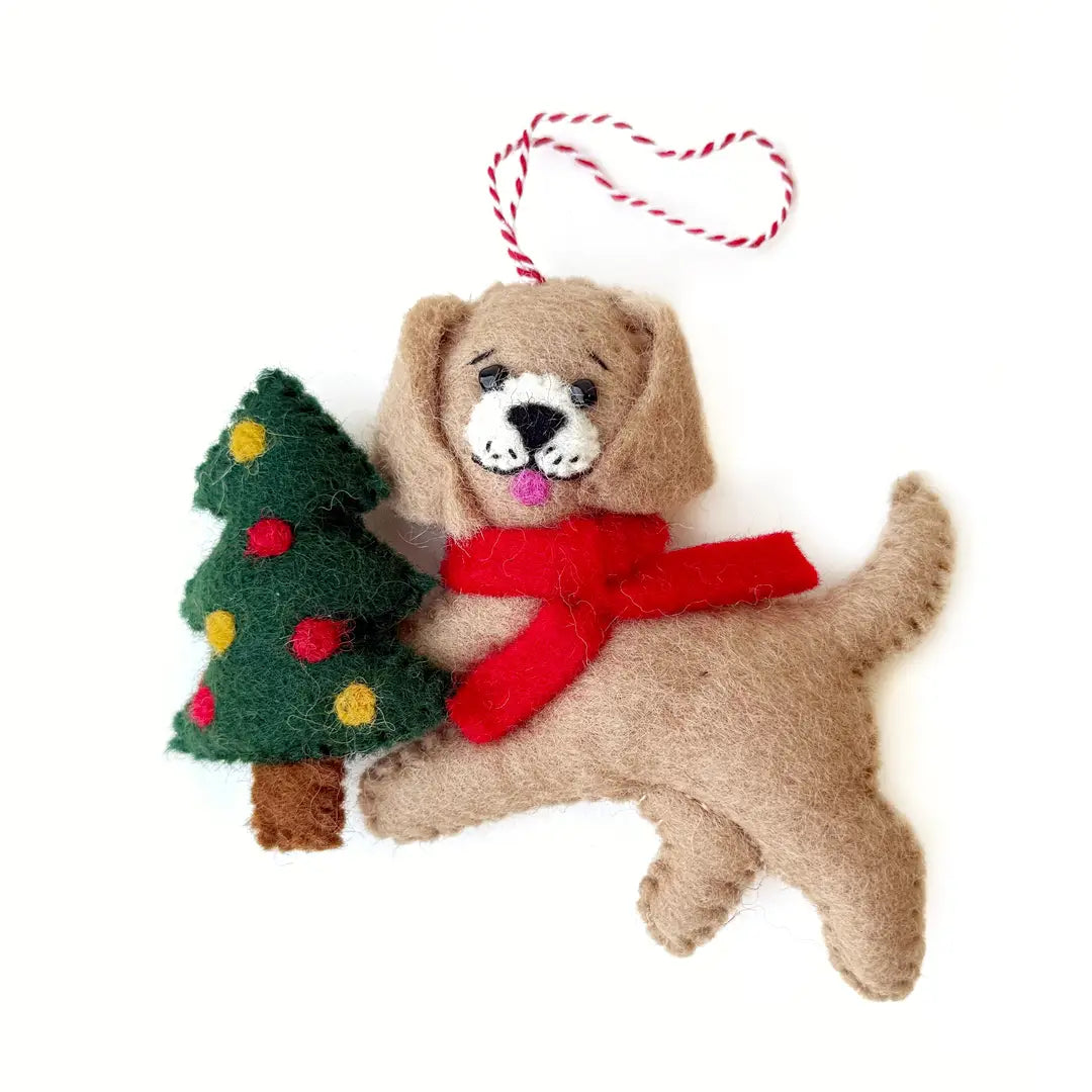 A felt Christmas ornament shaped like a tan puppy wearing a red scarf and holding a green Christmas tree with red and yellow ornaments, hanging from a red-and-white striped string on a white background.