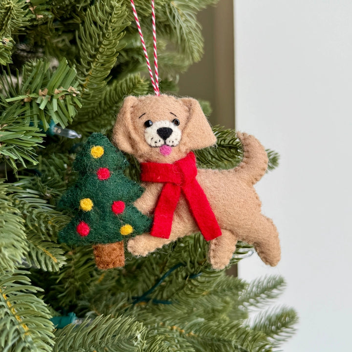 A felt Christmas ornament shaped like a tan dog wearing a red scarf, hanging on a green Christmas tree next to a small green felt tree with colorful ornaments.