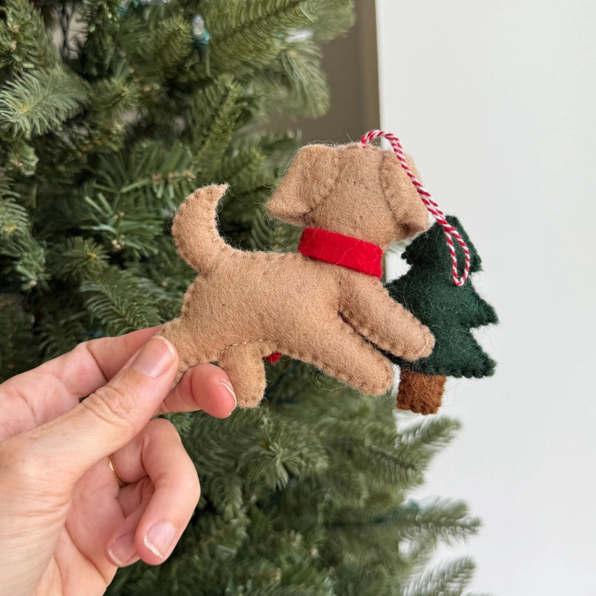 A hand holds a felt Christmas ornament shaped like a tan dog wearing a red bandana and carrying a dark green felt Christmas tree, with a blurred green Christmas tree in the background.