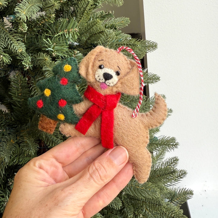A hand holds a felt Christmas ornament shaped like a tan puppy wearing a red scarf and holding a green Christmas tree, with a red-and-white hanging loop, in front of a real Christmas tree.