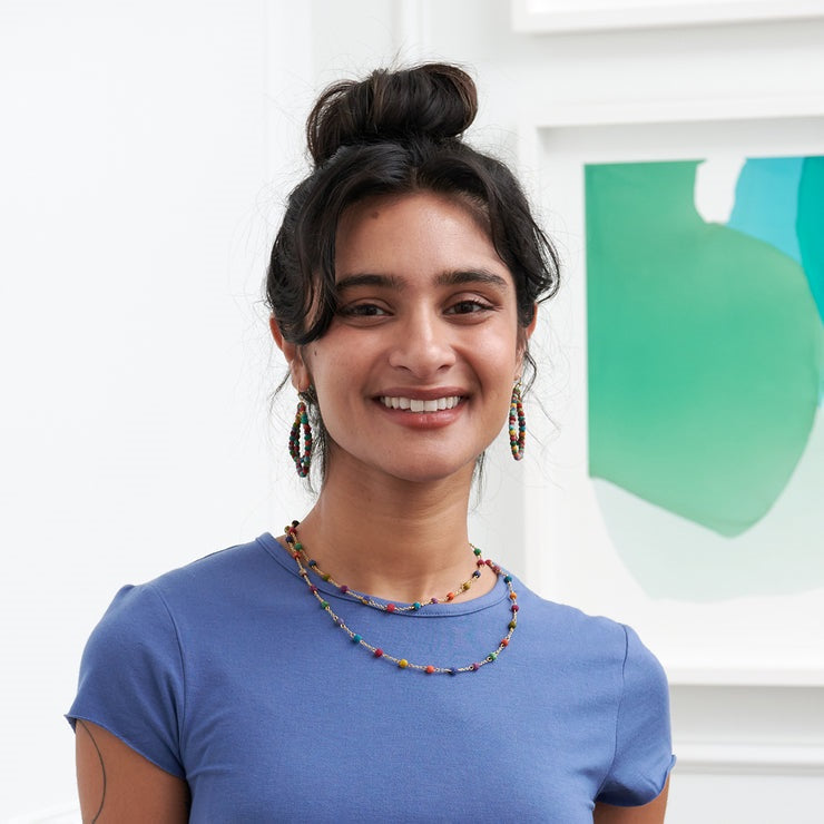 Woman wearing a blue shirt with colorful dotted necklace, standing in front of a white wall with a green abstract painting.