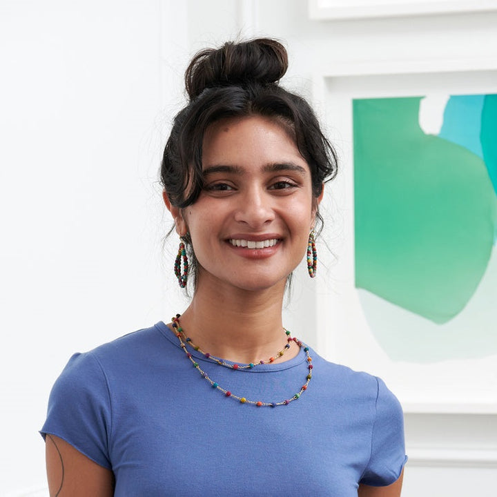 Woman wearing a blue shirt with colorful dotted necklace, standing in front of a white wall with a green abstract painting.