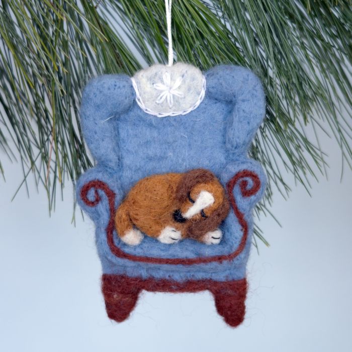 Decorative ornament of a dog on a blue chair hanging from a tree branch.