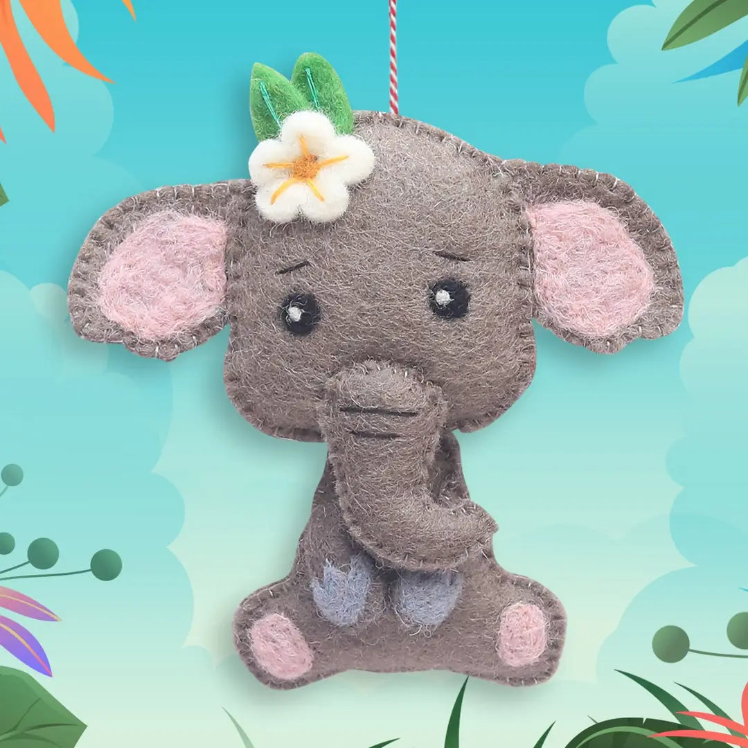 A felt elephant plush ornament hanging against a colorful tropical-style background. The elephant is gray with pink ears, pink and lavender feet, a white flower on its head, and a red-and-white hanging string.