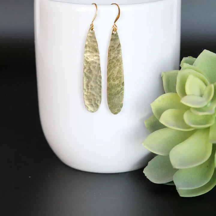 A pair of hammered olive green teardrop dangle earrings with gold fishhook wires, hanging on a white ceramic cup next to a green succulent on a black surface.