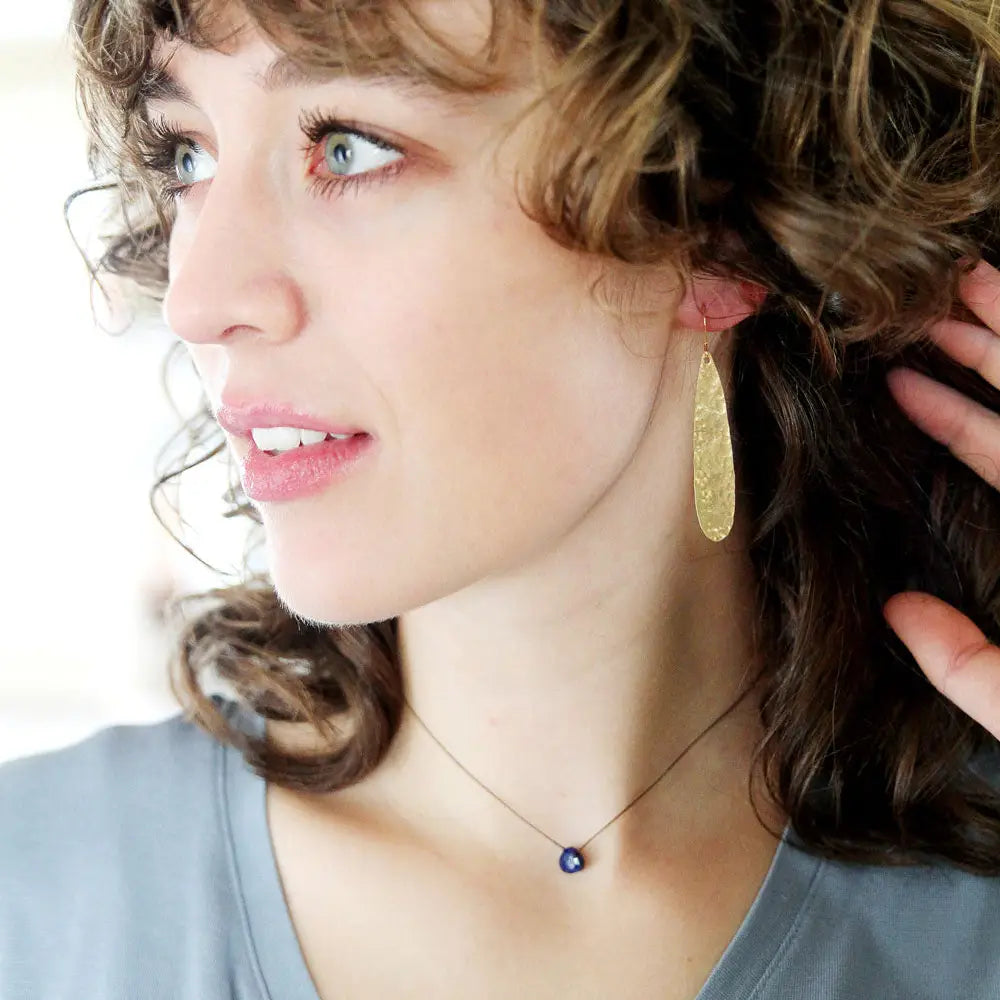 A close-up lifestyle shot of a woman with curly brown hair wearing a textured gold teardrop earring and a delicate necklace with a small dark blue bead.