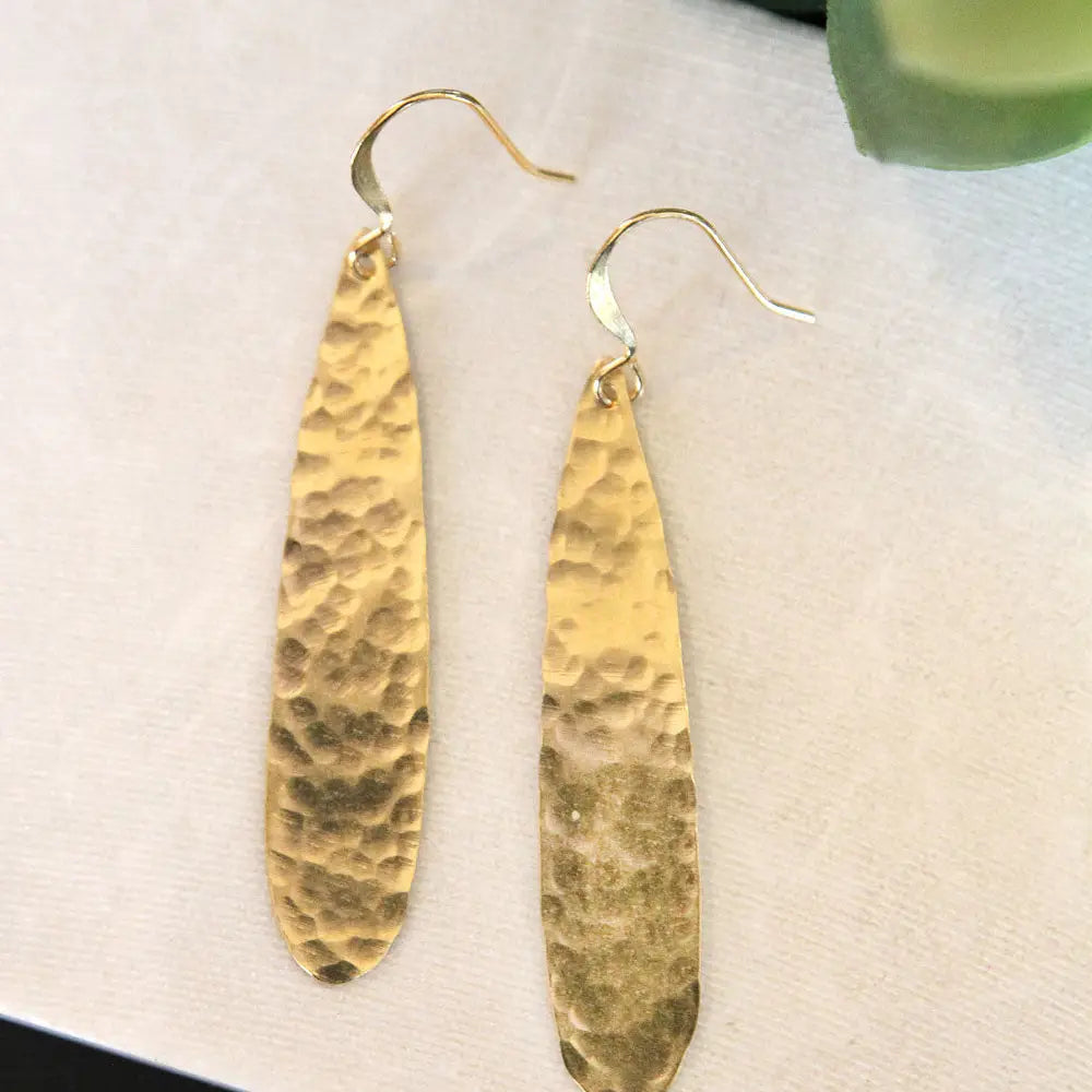 A pair of hammered gold dangle earrings with elongated teardrop-shaped plates and fishhook wires, displayed on a textured off-white surface.