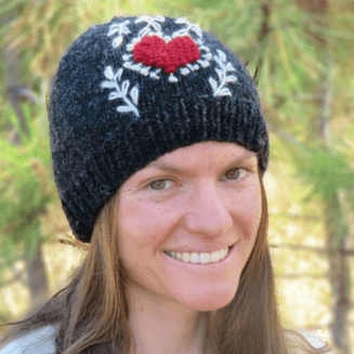 Person wearing a black knit beanie with red heart and white floral patterns in a forest setting