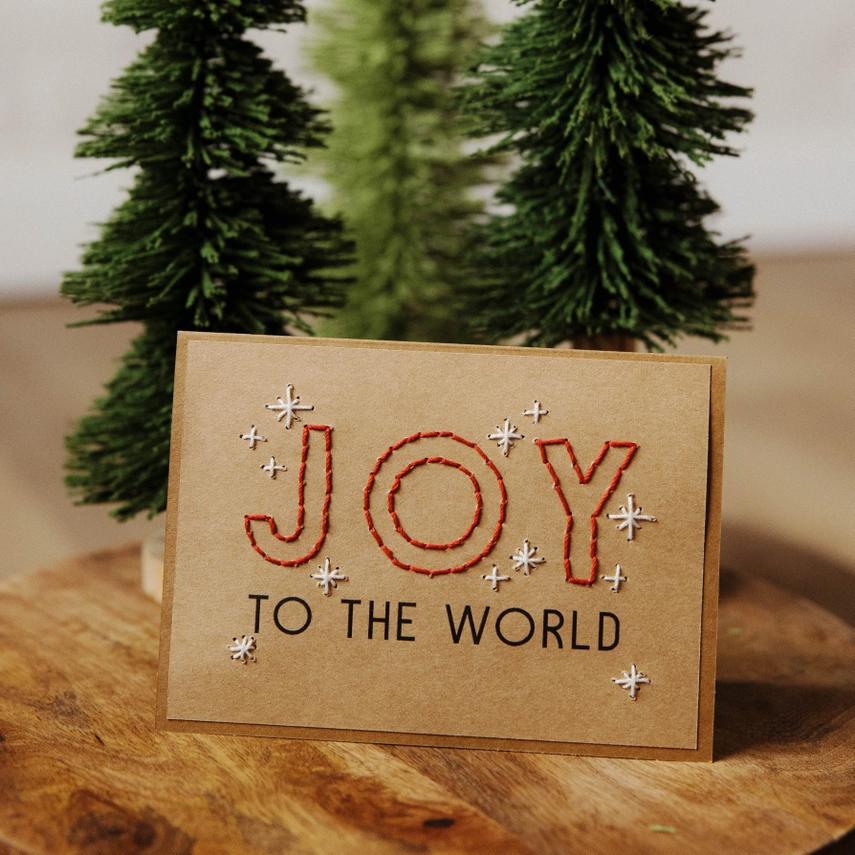 Card with 'Joy to the World' text on a wooden surface with Christmas trees in the background