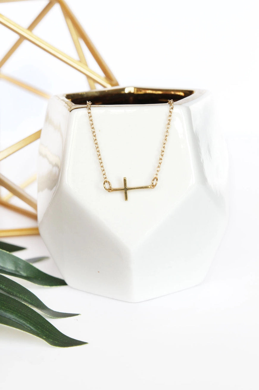 Gold necklace with a cross pendant on a white pot with gold interior, against a white background.