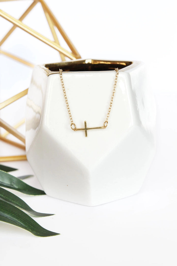Gold necklace with a cross pendant on a white pot with gold interior, against a white background.