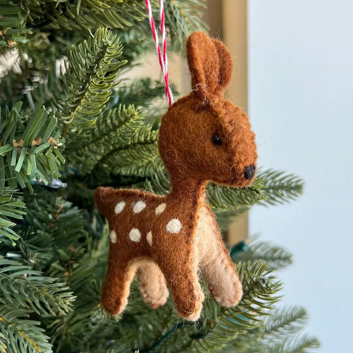 A brown felt deer ornament with white spots hanging from a red-and-white string on a green Christmas tree.