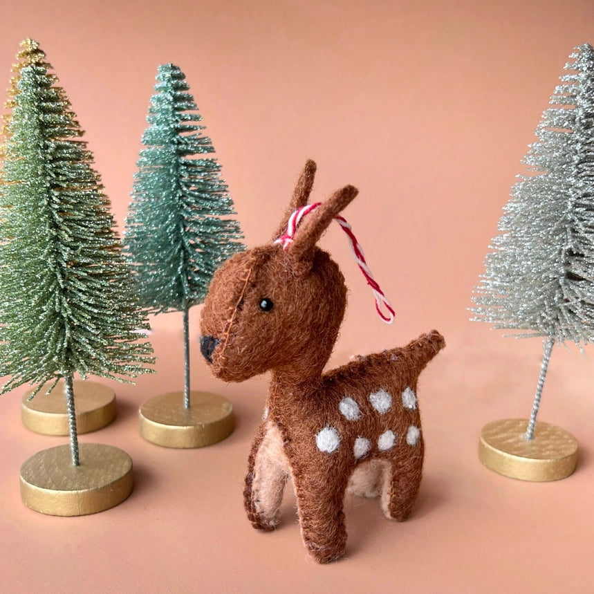 A small brown felt deer ornament with white spots and a red-and-white striped hanging ribbon, posed among decorative bottle-tree stands on a peach background.