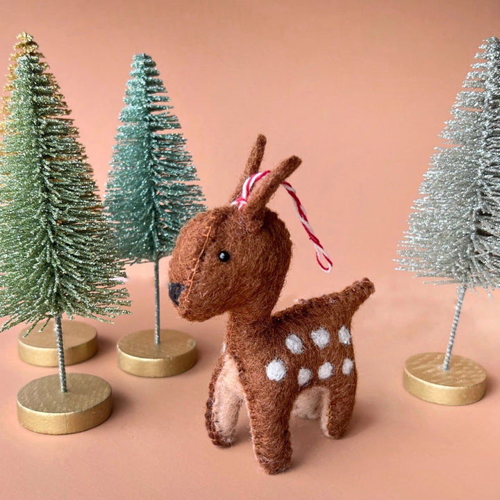 A small brown felt deer ornament with white spots and a red-and-white striped hanging ribbon, posed among decorative bottle-tree stands on a peach background.