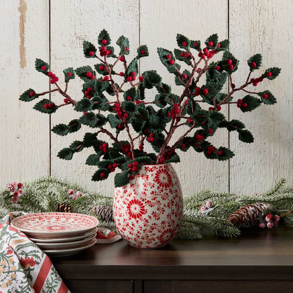 Decorative vase with red and white pattern containing holly branches on a wooden surface.