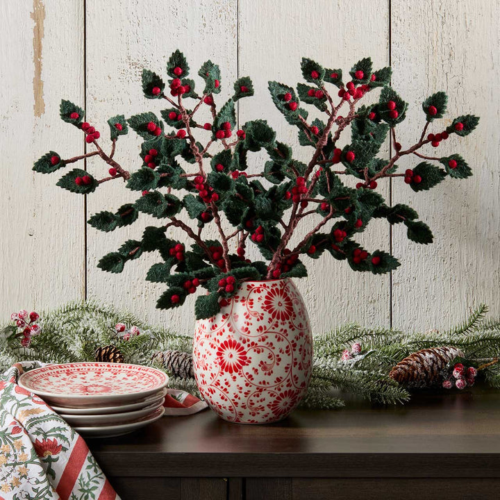 Decorative vase with red and white pattern containing holly branches on a wooden surface.