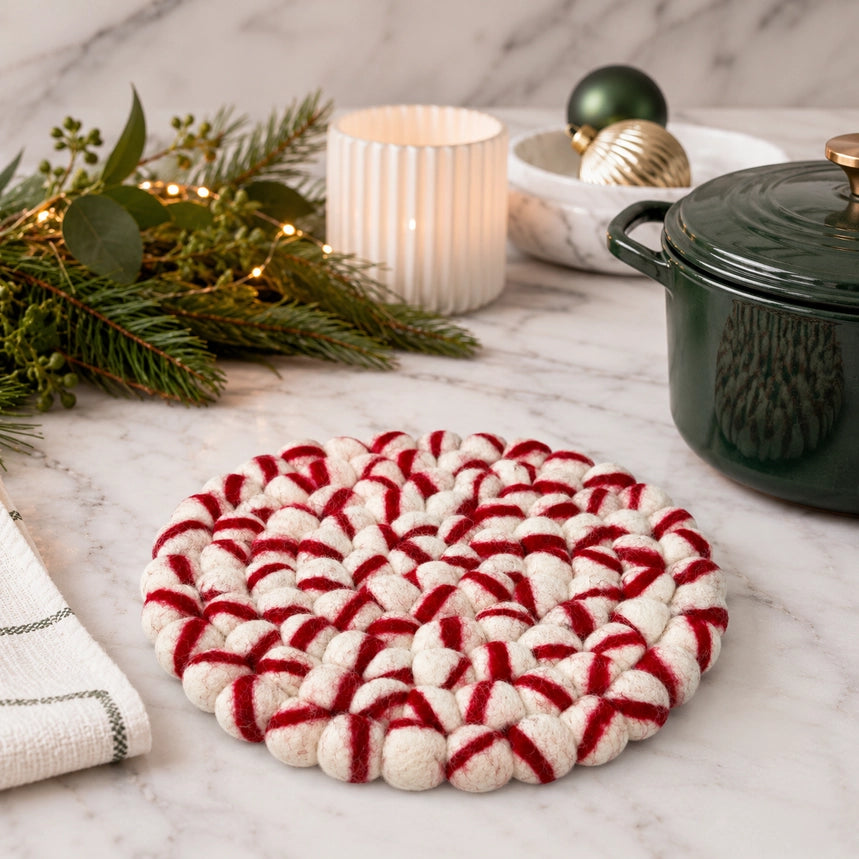 A round braided holiday trivet made of cream and red yarn, styled on a white marble surface with a green pot, candle, pine branches, and ornaments in the background.
