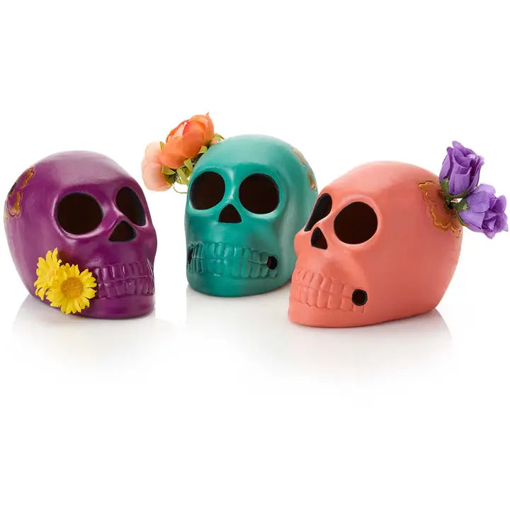 Three decorative skull-shaped candle holders in purple, teal, and coral pink, each styled with small artificial flowers, on a white background.