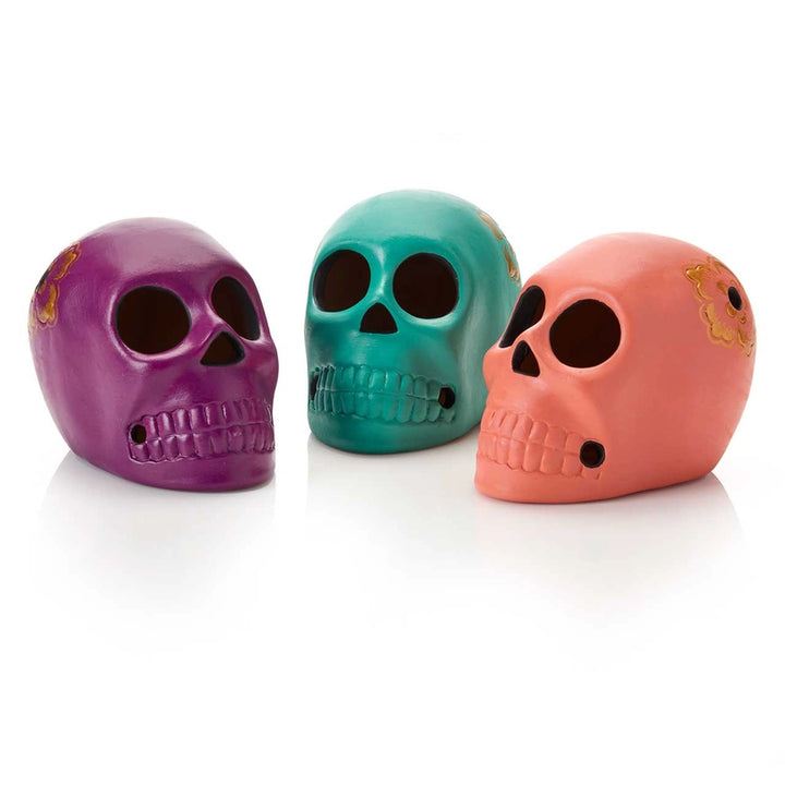 Three decorative skull figurines in purple, teal, and coral pink, arranged side by side on a white reflective surface.