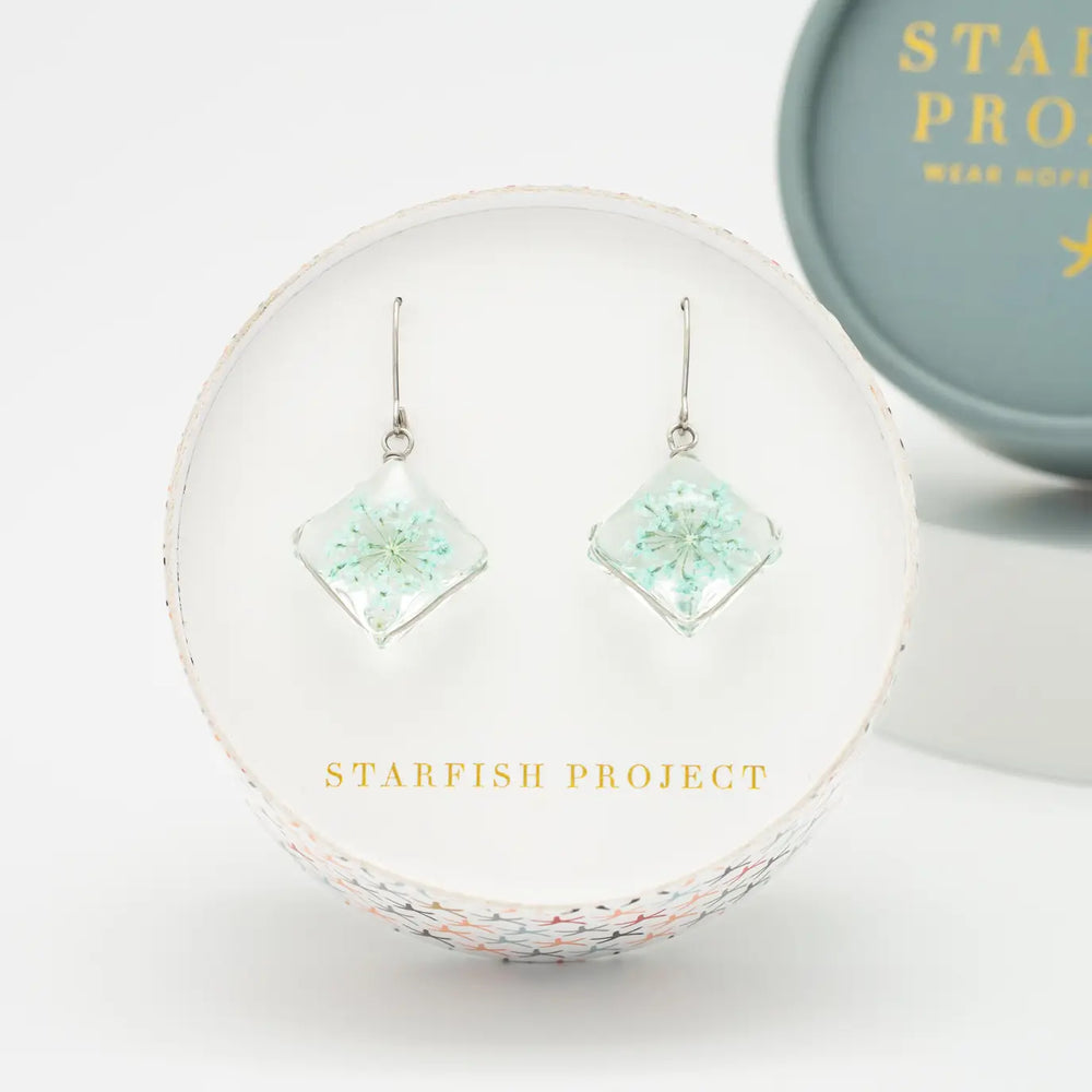 Pair of square earrings with floral design in a clear setting, displayed on a branded stand with 'Starfish Project' branding.