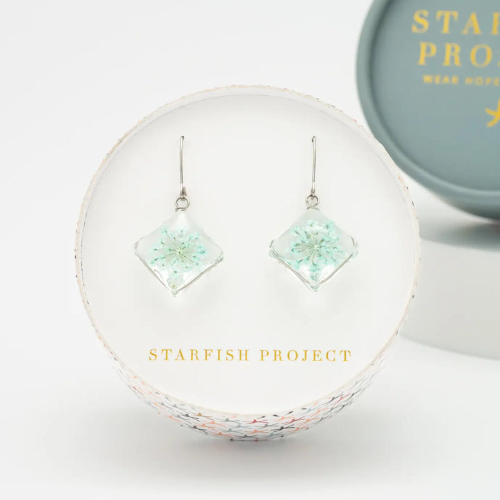Pair of square earrings with floral design in a clear setting, displayed on a branded stand with 'Starfish Project' branding.